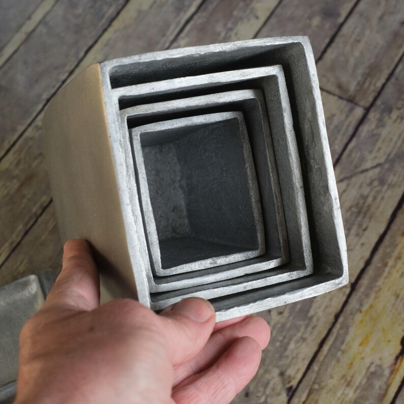 May include: A set of four square, nesting planters in varying sizes. The planters are made of a light grey metal with a slightly textured surface. The planters are stacked inside each other, showcasing their graduated sizes.