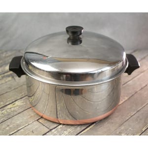 May include: A stainless steel pot with a lid and black handles. The pot has a copper-colored band around the base. The lid has a black knob. The pot is sitting on a wooden surface.