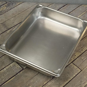 May include: A stainless steel rectangular food pan. The pan has a smooth, reflective surface and slightly rounded corners. The pan is empty and sits on a wooden surface.