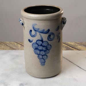 May include: A stoneware crock with a blue and white grape design. The crock has two handles and a black glazed interior.