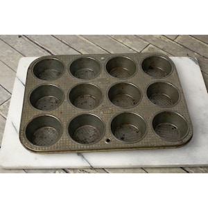 May include: A vintage metal muffin tin with 12 individual cups. The tin is gray and has a textured surface.