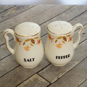 May include: A pair of cream-colored ceramic salt and pepper shakers with handles. Each shaker features a gold-trimmed rim, a floral pattern with brown, orange, and yellow leaves and flowers, and the words SALT or PEPPER printed in black.