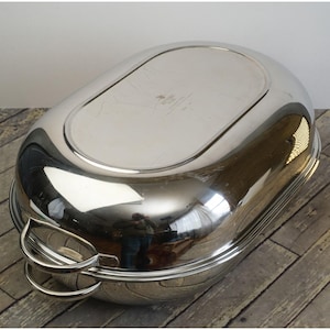May include: A shiny, oval-shaped, stainless steel serving dish with two curved handles. The dish has a reflective surface, and the interior is a lighter shade of silver. The dish is sitting on a wooden surface.