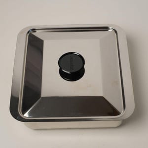 May include: A stainless steel square baking dish with a black plastic lid. The lid has a small knob on top with the word "West Bend" printed on it.