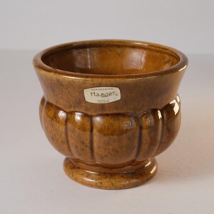 May include: A brown ceramic planter with a textured, ribbed design. The planter has a wide opening and a rounded base. A white sticker with the word "Haeger" is on the front. The planter is likely vintage.