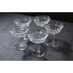 May include: Five clear crystal champagne coupe glasses. Each glass features a round bowl with a diamond pattern and a slender stem. The glasses are arranged on a dark gray surface.