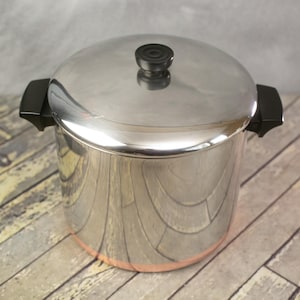 May include: A stainless steel stockpot with a lid and black handles. The pot has a copper-colored base and a black knob on the lid. The pot is designed for cooking and food preparation.