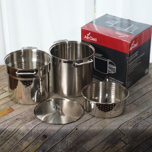 May include: A stainless steel All-Clad multi-cooker set including a large stockpot, a pasta insert, a steamer basket, and a lid. The set is displayed next to its original red and black cardboard packaging box with the All-Clad logo printed on it.