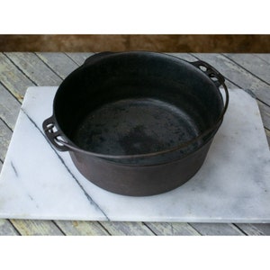 May include: A black cast iron Dutch oven with two handles on the side and a wire bail handle on top. The oven is sitting on a white marble surface.