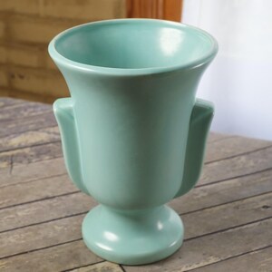 May include: A light teal ceramic vase with a flared top and a rounded base. The vase has two decorative handles on the sides and stands approximately 7 inches tall. The vase is a solid color and has a glossy finish.