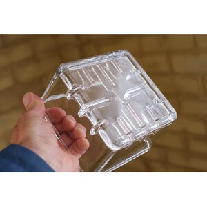 Pu&ograve; includere: Un contenitore di plastica trasparente di forma quadrata con coperchio. Il contenitore ha una superficie strutturata ed &egrave; tenuto da una persona. Il coperchio presenta creste e scanalature in rilievo. Per la conservazione degli alimenti.