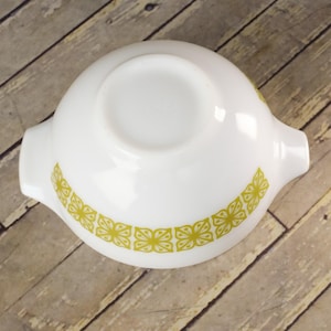 May include: White Pyrex bowl with two handles and a decorative band of yellow-green floral designs. The bowl is round and has a smooth, glossy finish. The bowl is set against a wooden background.