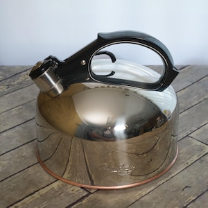 May include: A polished stainless steel tea kettle with a reflective, rounded body and a copper-toned base. It features a black plastic handle with an integrated trigger mechanism and a metallic spout cap. The brand name is etched on the side.
