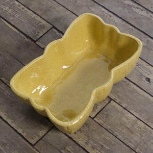 May include: A yellow ceramic bowl with a scalloped, wavy rim and a glossy, speckled finish. The bowl has an elongated, irregular shape with rounded edges and a smooth, reflective interior surface, showcasing a vibrant mustard yellow hue.
