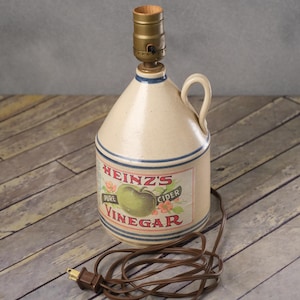 May include: A vintage Heinz vinegar jug repurposed as a lamp. The beige ceramic jug features a label with "Heinz's Pure Cider Vinegar" text and a green apple illustration. It has a handle and is accented with blue stripes. A brown electrical cord and lamp fixture are attached.
