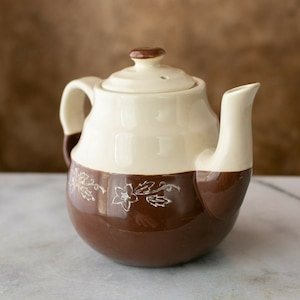 May include: A brown and cream ceramic teapot with a floral design. The teapot has a lid and a spout.