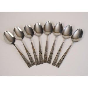 May include: A set of eight stainless steel spoons arranged in a fan shape. The spoons have oval bowls and long, slender handles with an ornate, engraved design near the base. The spoons are reflective, with a bright, metallic sheen.
