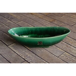 May include: A green ceramic oval serving bowl. The bowl has a glossy finish and a slightly textured surface. The label on the bowl reads "Le Mar Imperial". The bowl is placed on a wooden surface.