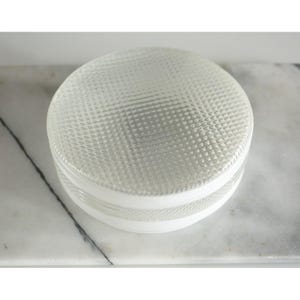 May include: A clear, round glass object with a textured, diamond-patterned top and a white band around the middle. The object is sitting on a white and gray marble surface. The object appears to be a decorative container.