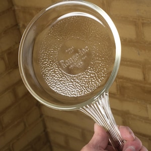 May include: Clear glass skillet with a handle. The skillet has a textured bottom and the words "Range-tec" are visible. The handle is ribbed and the skillet is held up against a brick wall.