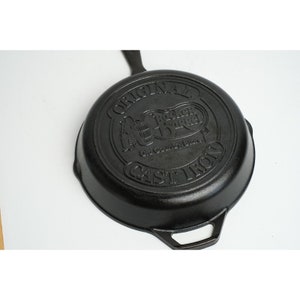 May include: Black cast iron skillet with a raised logo that reads "Original  Rucker Barrel Old Country Store Cast Iron".