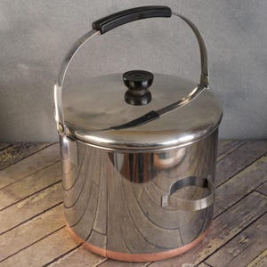 May include: A stainless steel ice bucket with a lid and a black handle. The bucket has a copper-colored base and a silver handle. The lid has a black knob. The bucket has a handle on the side. The bucket is sitting on a wooden surface.