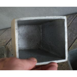 May include: A close-up of a square, metallic planter with a textured, grey interior and a lighter, off-white exterior. The planter has a simple, geometric design, and the edges are slightly rounded.