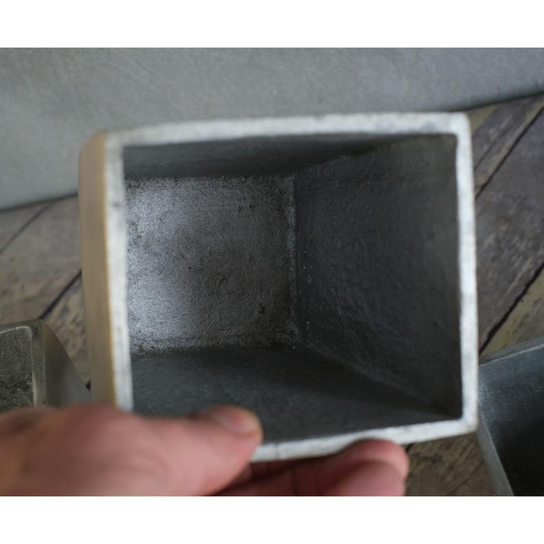 May include: A close-up of a square, metallic planter with a textured, grey interior and a lighter, off-white exterior. The planter has a simple, geometric design, and the edges are slightly rounded.
