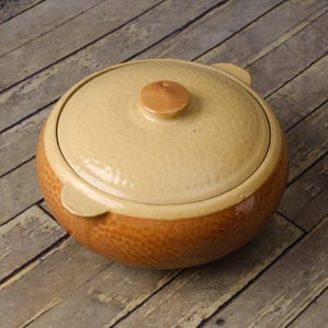 May include: A round, lidded ceramic casserole dish. The dish is primarily beige with a textured, speckled pattern on the lower half and lid. It has a brown band around the base and a small handle on each side.