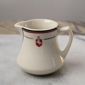 May include: A small, white ceramic creamer with a black and burgundy trim. The creamer features a red crest with a white horse and the letters "R" and "R" on a white shield.