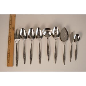 May include: A collection of stainless steel silverware, including forks, spoons, a ladle, a cake server, a knife, and a teaspoon. Each piece has a floral design on the handle. A wooden ruler is on the left side.