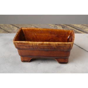 May include: A brown ceramic planter with a rectangular shape and four small feet. The planter has a glazed finish and a decorative pattern around the top edge.