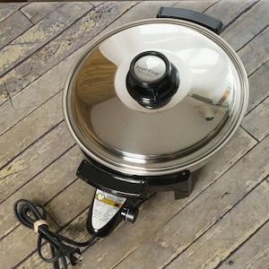 May include: A round electric skillet with a polished stainless steel finish and a matching lid. The lid features a black handle with a heart logo and the text Lustre Craft. A black control base includes a power cord and a label with text.