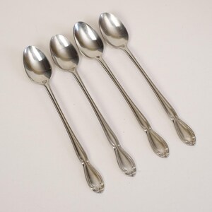 May include: Four stainless steel spoons with long, slender handles and oval bowls. The handles have a decorative, symmetrical design near the base. The spoons are arranged diagonally against a white background, reflecting light.