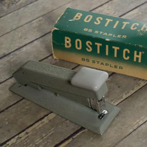 May include: A vintage Bostitch B5 stapler with a textured olive-gray finish and a smooth, light gray top press plate. The stapler is positioned next to its original rectangular green and tan cardboard box, which features the text BOSTITCH B5 STAPLER.