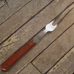 May include: A two-pronged carving fork featuring a polished stainless steel head and a wooden handle secured with three metal rivets. The metal neck of the fork displays an etched inscription that reads "STAINLESS STEEL" in a clear font.