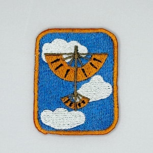 May include: A rectangular embroidered patch featuring a blue sky background with white clouds. The central design depicts a vertical structure with two orange, fan-shaped wings with black line details, all enclosed by a thick orange border.