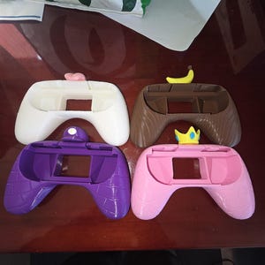 May include: Four brightly coloured game controller shells. The shells are white with a pink heart, brown with a banana, purple with a brick pattern, and pink with a gold crown. The controllers are designed to fit a gaming device.