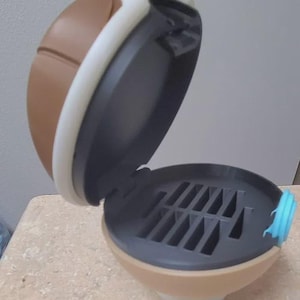 May include: An open, brown, and tan container with a black interior. The container has a hinged lid with a white trim. The interior has multiple slots. A blue tab is on the side of the container.