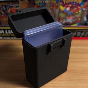 May include: A black card storage box with a hinged lid, open to show clear card sleeves. The box has a handle and is on a wooden surface. The box is designed for storing trading cards.