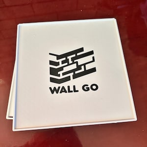May include: A square white tile with a black graphic of a stylized brick wall corner and the text WALL GO printed in black capital letters. The tile has a thin white border and a smooth, flat surface, presenting a clean and simple design.