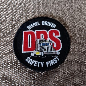 May include: A round black patch with the text "DIESEL DRIVER" above large red letters "DPS" and a truck graphic. Below the letters is the text "SAFETY FIRST" in white. The patch is likely for a truck driver.