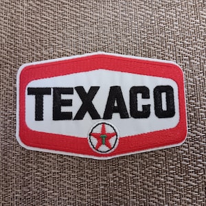 May include: Embroidered Texaco patch with a red and white border. The patch features the word "TEXACO" in bold black letters on a white background. A small circular emblem with a red star is at the bottom.