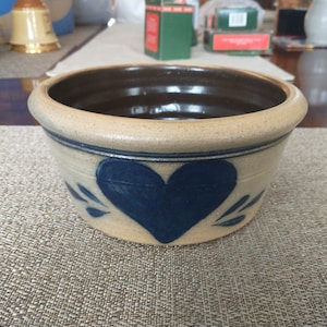May include: A small, beige ceramic bowl with a blue heart design. The bowl has a brown glaze on the inside and a white glaze on the outside.