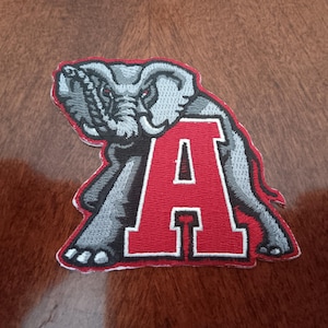 May include: Embroidered patch featuring a gray elephant with red eyes and a red letter "A". The elephant is positioned over the letter "A", which has a white outline. The patch has a white border.