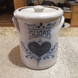 May include: A white ceramic sugar jar with a blue heart and floral design. The jar has a lid and two handles. The word "SUGAR" is written on the front of the jar.