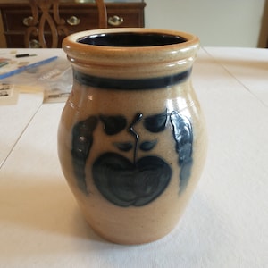 May include: A brown ceramic vase with a blue glaze and a painted apple design. The vase has a wide mouth and a rounded body.