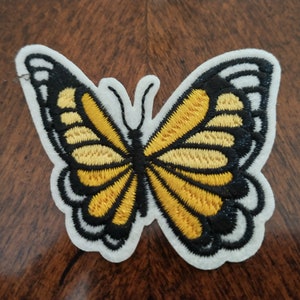 May include: Embroidered butterfly patch with a white border. The butterfly features yellow and black wings with detailed stitching. The patch is designed to be sewn or ironed onto fabric, suitable for clothing or accessories.