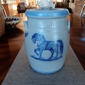 Puede incluir: Un tarro de gres antiguo con un diseño de caballo azul y blanco. El tarro con tapa tiene un borde y asas de color azul claro. El caballo está representado en una ilustración estilizada detallada. El año "1892" está impreso en el tarro.