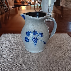 May include: A stoneware pitcher with a gray speckled finish and a dark blue interior. The pitcher features a blue floral design on the front and a curved handle. It is sitting on a textured placemat.